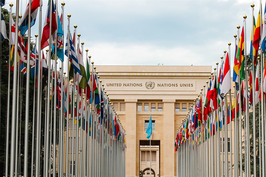 [ai] The building of the United Nations in Geneva, viewed from a pathway lined with flags of various countries, under a cloudy sky.